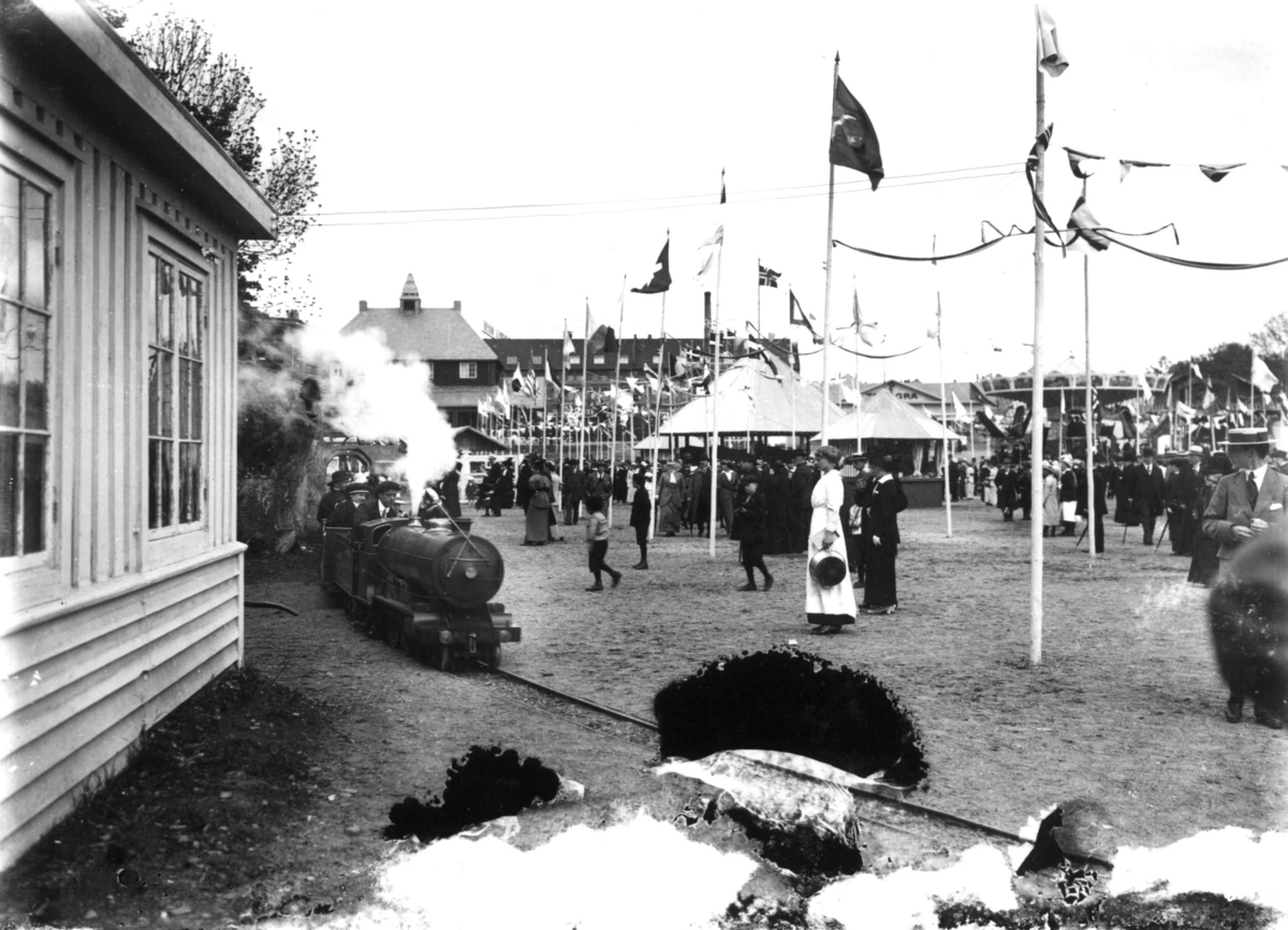 Jubileumsutstillingen på Frogner, Oslo, 1914.
Lilleputtbane. Publikum og paviljonger i bakgrunnen. Flagg.