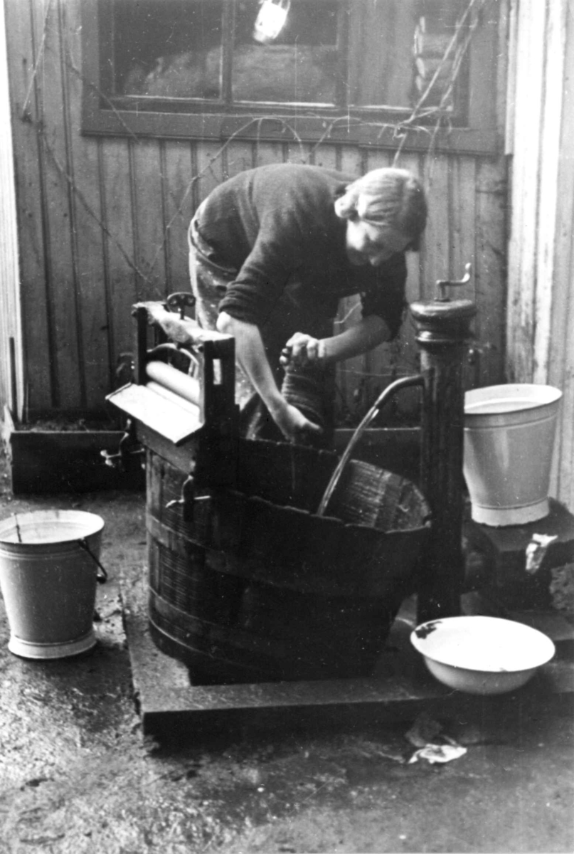 Klesvask, kvinne vasker tøy utendørs, utstyrt med vannpost, rulle/maskin til å presse ut vann, emaljerte bøtter og fat. Jfr. NF.13400-073. Fra boliginspektør Nanna Brochs boligundersøkelser i Oslo 1920-årene.