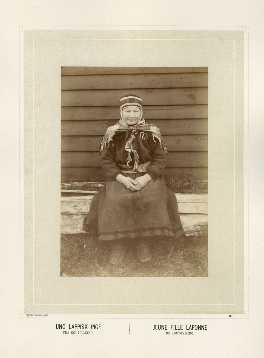 Ung samisk kvinne med kofte og sjal sitter på en benk utenfor bygning.