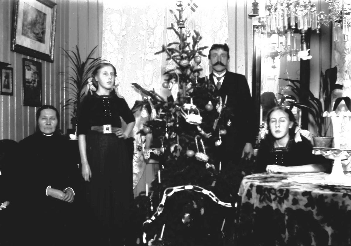 Stueinteriør. Julefeiring. Mann og tre kvinner, pyntede juletreet, bord, stoler, lampe, gardiner. Oslo, ca 1915