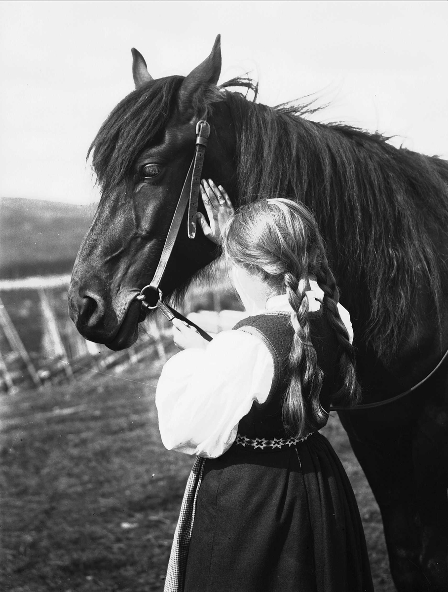 Jente i nasjonaldrakt holder i tømmene til en hest og klapper den på nakken. Gjerde i bakgrunnen.