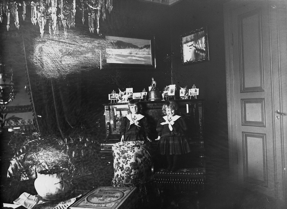 Karen og Gudrun Q. Wiborg sitter på piano i stue, Meltzers gate 9, Oslo, 1899. Fotografier i ramme plassert på pianoet.