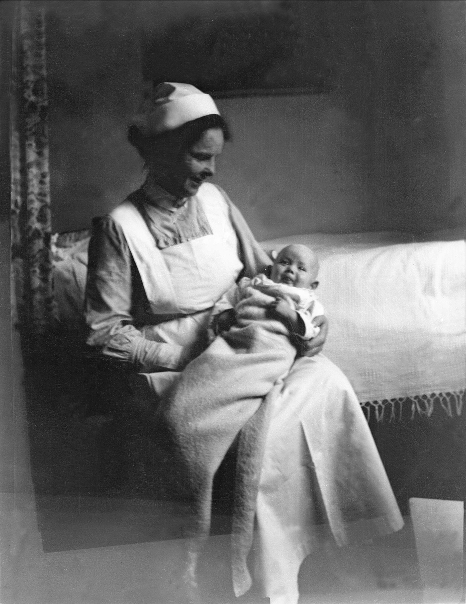 En kvinne sitter med en baby på sitt fang ved en seng. Robsahm og Lund.
