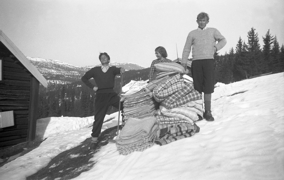 To unge menn og en jente, ant. Thode Holm, Per Klem og Kari Arentz, ryster tepper og dyner ved Arentz-familiens feriebolig Ligardshaugen I Eggedal. Fotografert påsken 1948.