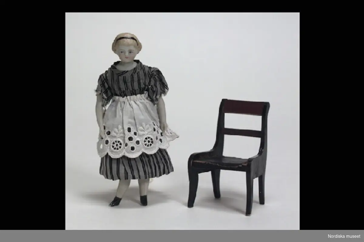 Inventering Sesam 1996-1999:
Docka  H  13    Stol  H  7,5    Barnbädd  L  9,5   B  6 (cm)
Dockskåpsdocka, kvinna, sittande (fastnålad) på stol, med barnbädd och babydocka (a-c). 
a) Docka; huvud, sköld, halvarmar och halvben av biskvi, kropp i stoppat tyg, målade ansiktsdrag och blont hår, svart diadem, grå sidenklänning med svarta ränder, vitt förkläde, underkjol och mamelucker, svartmålade kängor modellerade i massan. 
b) Stol, av svartmålat lövträ, rygg med brunbetsat överstycke och tvärslå, något svängda ben. Under bottnen med blyerts. "Thyra".
c) Stoppad bädd, klädd med vitt bomullstyg, garnerad med spets och rosa sidenband, fastsydd babydocka av biskvi, iklädd rosa dräkt med spetskrage.
Tillhör dockskåp från omkr 1840, inv nr 145.505, inrett 1872 av leksakshandlare W A Bandel, Västerlånggatan, Stockholm, för dottern Thyra, vilken vid överlämnandet till museet själv gjort uppställningen av föremålen i dockskåpet. 
Anna Womack dec 1997