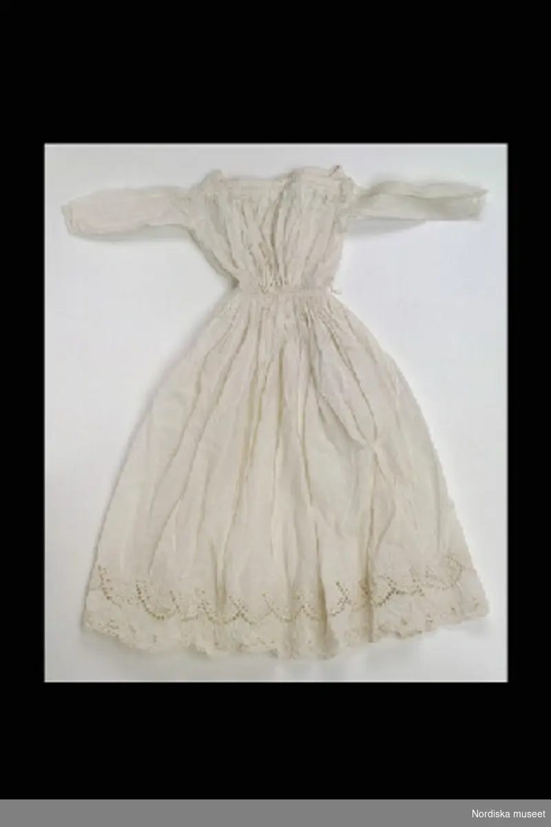 Inventering Sesam 1996-1999:
L 47 (cm)
Klänning, dockklänning, av vitt bomullstyg (linong), rak halsringning, lång ärm, vid rynkad kjol med dragsko i midjan och brodyr nedtill. 
Anna Womack jan. 1997