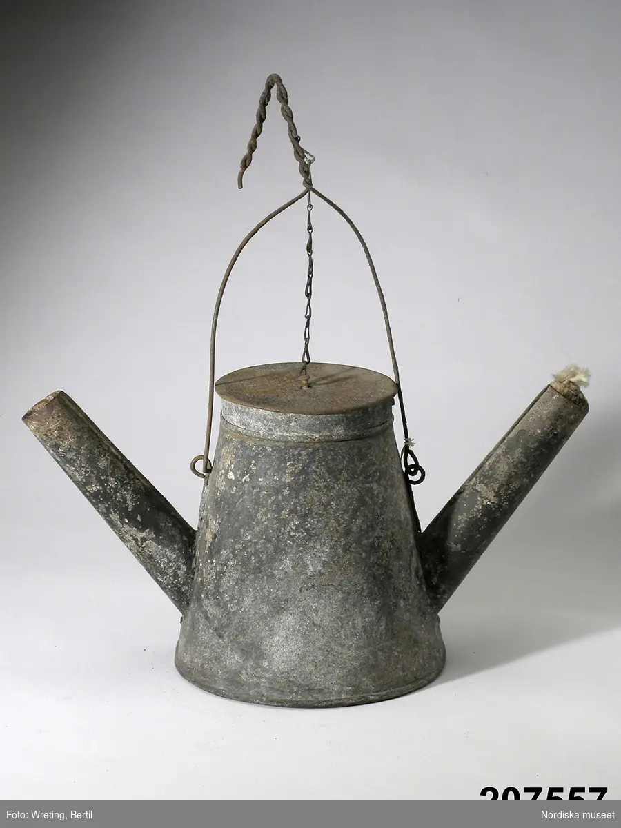 Blosslampa, dubbelpipig, bleckplåt, 18-1900-tal.
Konande kanna med två pipar, lock och ett bärhandtag.
I varje pipöppning sticker det fram en härva av bomullstråd (veke). 
Drivs med fotogen.