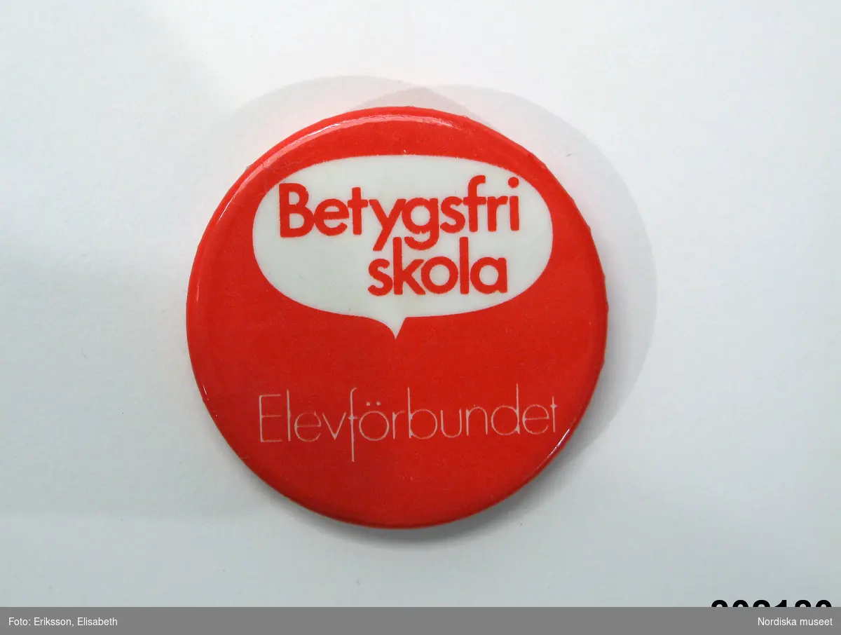 Rund knapp av bleckplåt överdragen med plastskikt i rött och vitt med texten "Betygsfri skola" i en pratbubbla och därunder: "Elevförbundet". Nål och krylle på baksidan.
/Berit Eldvik 2009-12-18