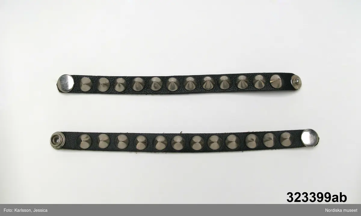 Två armband i svart läder med nitar i vitmetall längs hela bandet. Inköpta i punkaffär i Brighton c. 1980-81.