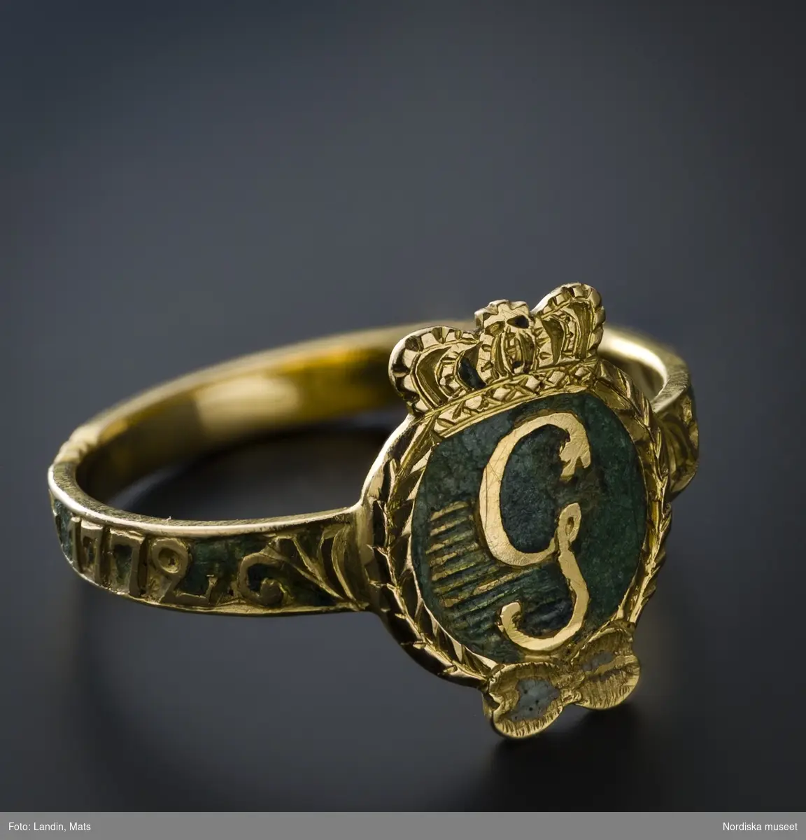 Ring av rött guld,  med Gustav III:s monogram i guld mot grön emaljbotten, s.k. revolutionsring. Runt skenan graverad inskription "den 19 aug 1772". 
/Ingrid Roos 2006-09-26