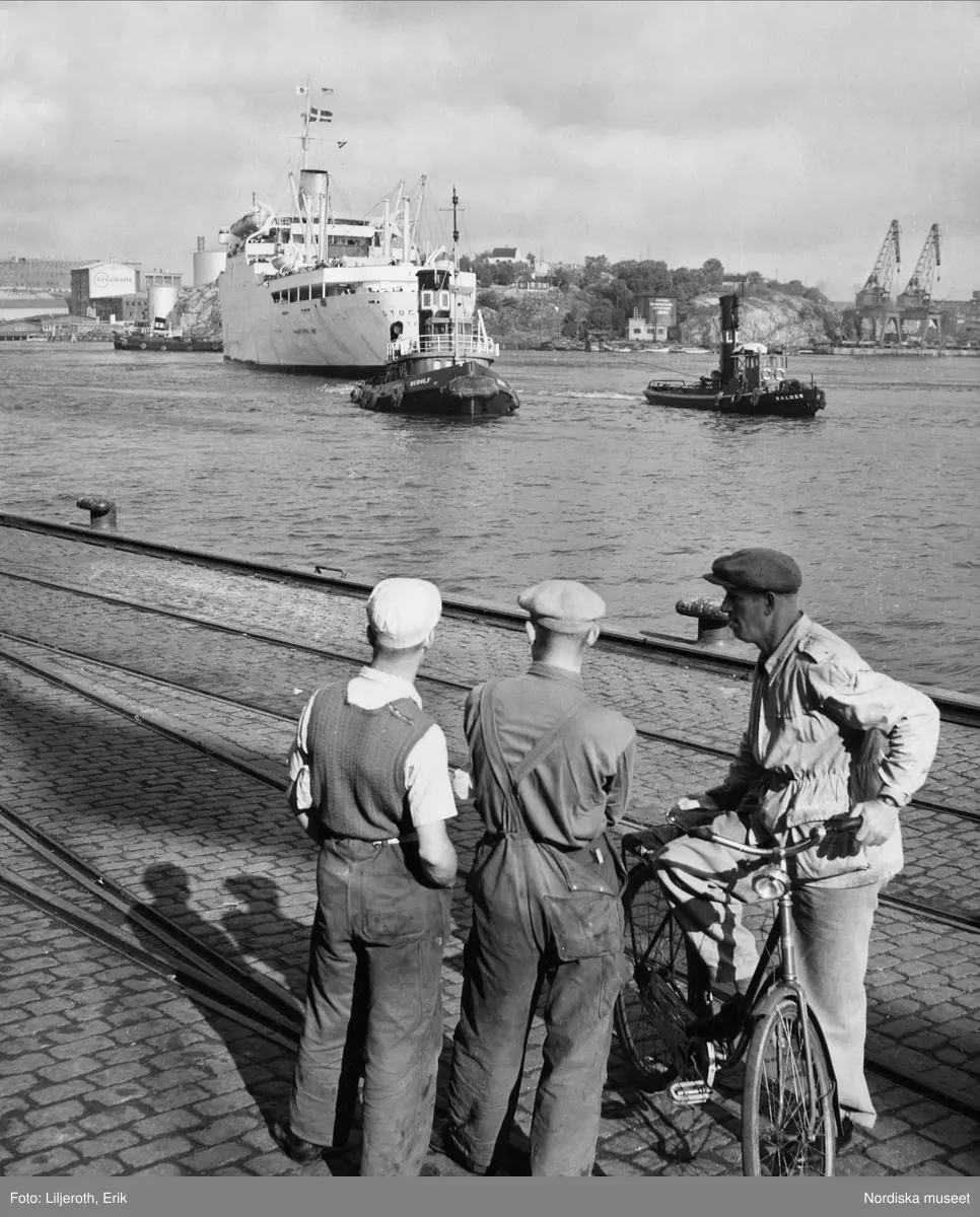 Tre hamnarbetare tittar på Svenska Amerika Liniens passagerarfartyg M/S Stockholm på väg in i Göteborgs hamn