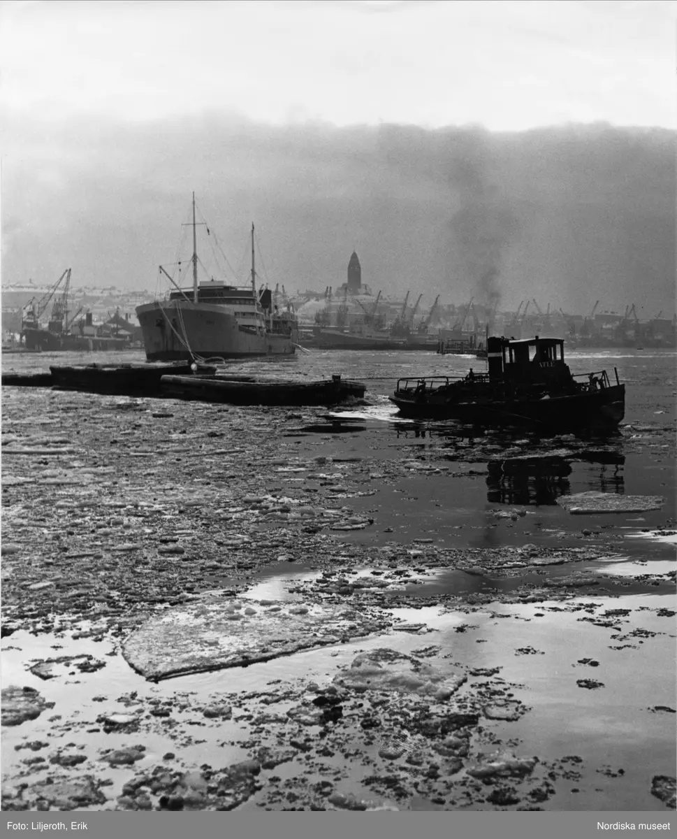 I förgrunden en bogserbåt och ett fraktfartyg. I bakrunden syns silhuetten av Masthuggsbebyggelsen