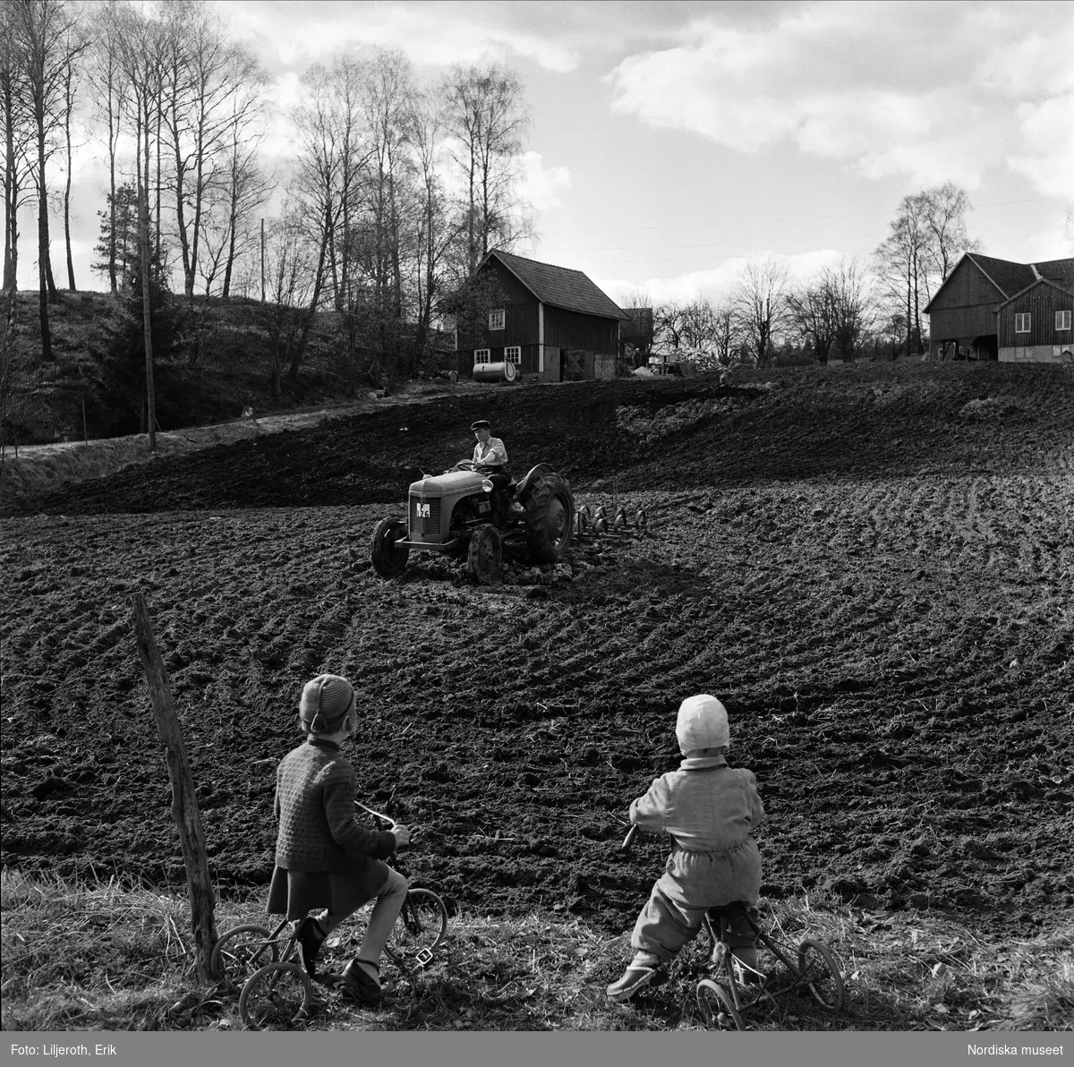 Vårbruk på en småländsk åker. En lantbrukare harvar med traktor (c-pinnharv). I förgrunden två små barn på varsin trehjuling.