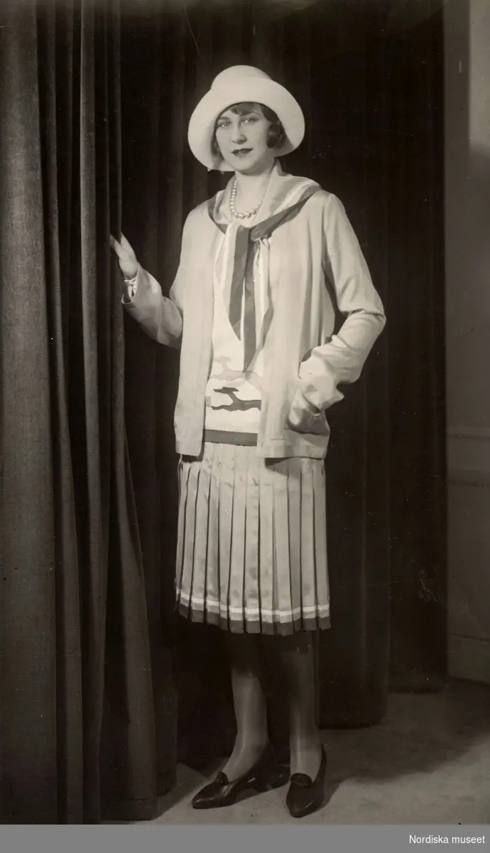 Modevisning i mars 1929. Kvinna iklädd dräkt med plisserad kjol och hatt. Nordiska Kompaniet.