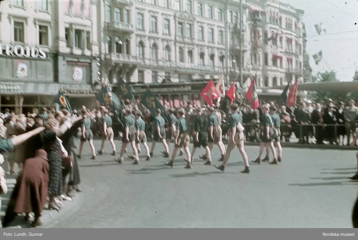 Politisk demonstration av ett nazistiskt parti vid Stureplan, Stockholm. Ungdomar, pojkar, i gröna skjortor och kortbyxor bär fanor.