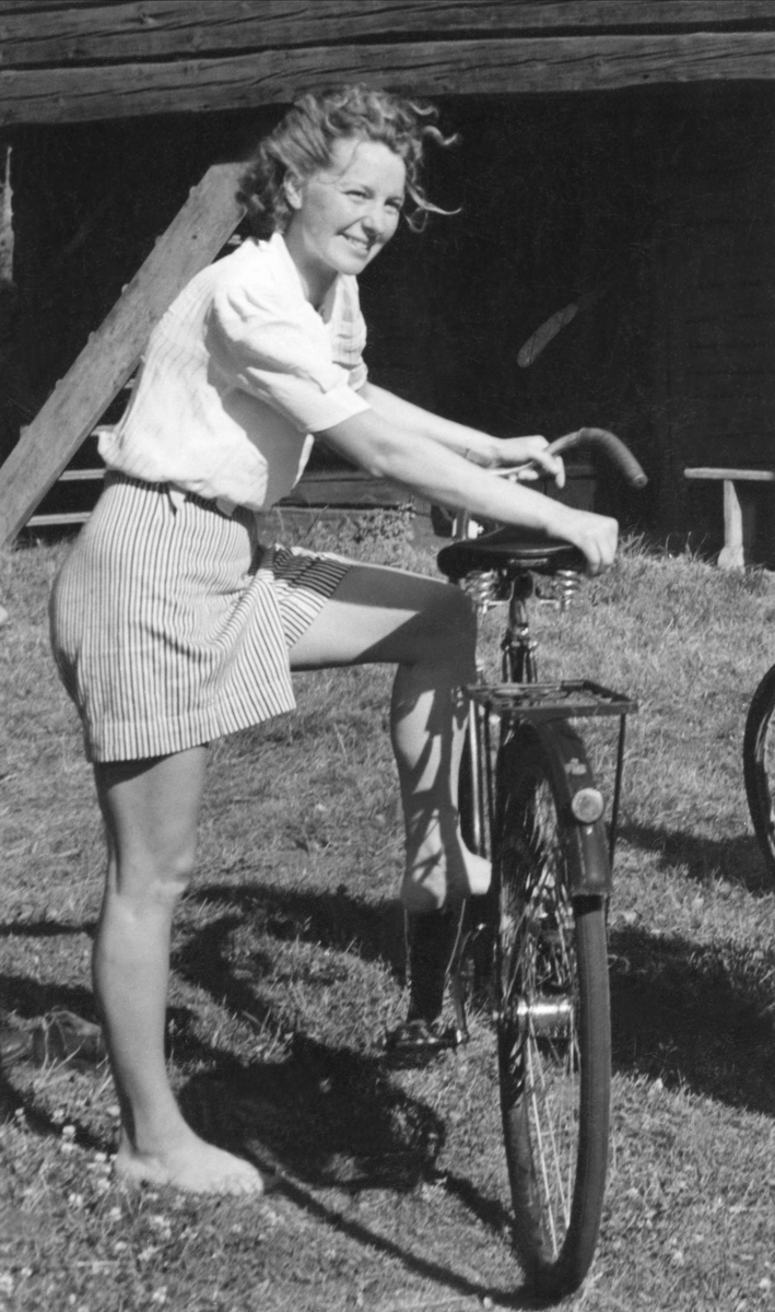 Flicka poserar vid cykel. Hon är klädd i kortärmad blus och randiga kortbyxor.