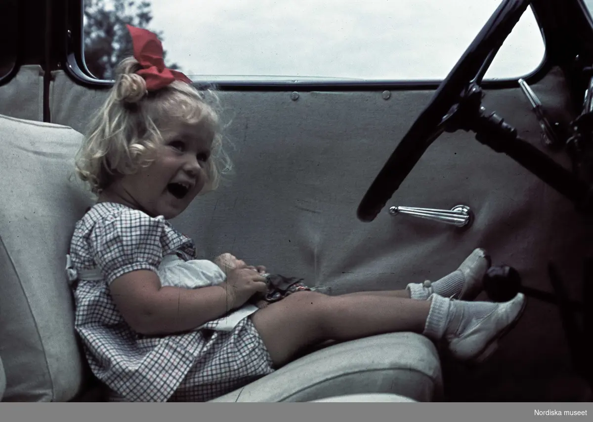 En liten flicka med röd rosett i håret sitter på förarplatsen i en bil. I knät håller hon en docka.