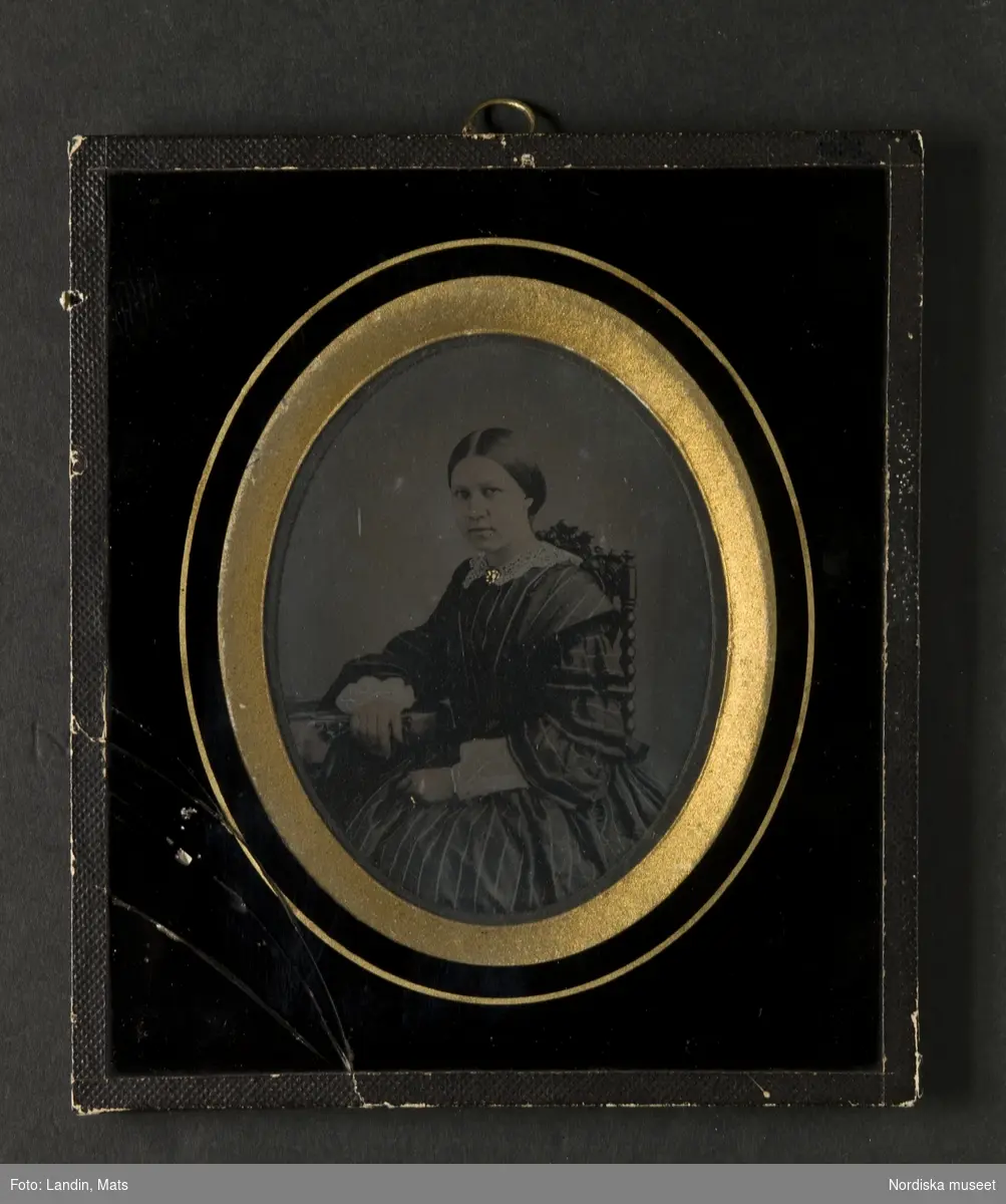 Porträtt, höftbild, av ung kvinna. Bilden föreställer fru Augusta Flack, född Frestadius (1832-1912). Sitter i stol med högt ryggstycke, klädd i mörkrandig klänning med vida ärmar och volanger samt vit krage. Mittbenat uppsatt hår. Brosch och ring förgyllda. Ambrotyp i glasad ram. På bakstycket påklistrad vit etikett med svart tryckt text: "W.A. Eurenius & P.L. Quist/FOTOGRAFISK ATELIER/Stockholm/Regeringsgatan No 14". Med bläck årtalet "1858".
Nordiska museet inv.nr 117638
