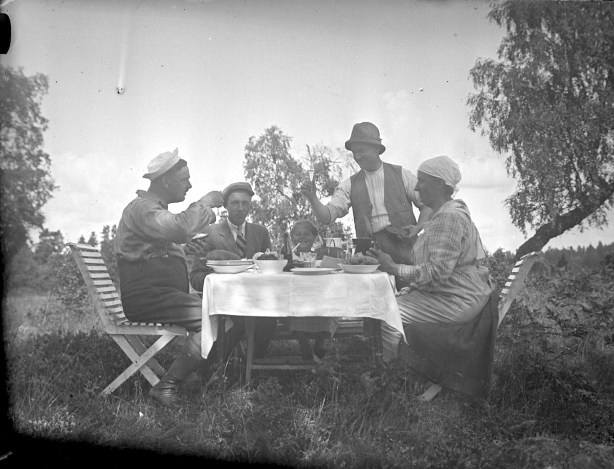 Tre män, en kvinna och en flicka kring ett dukat middagsbord utomhus. Två av männen höjer sina nubbeglas och skålar.