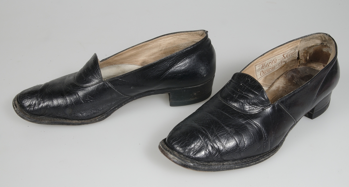 Ett par skor av svart skinn. Halvsulad lädersula. Fodrad med brunt skinn och lärft. Märkt: Marta-skor Oscaria-Örebro. Storlek 38.