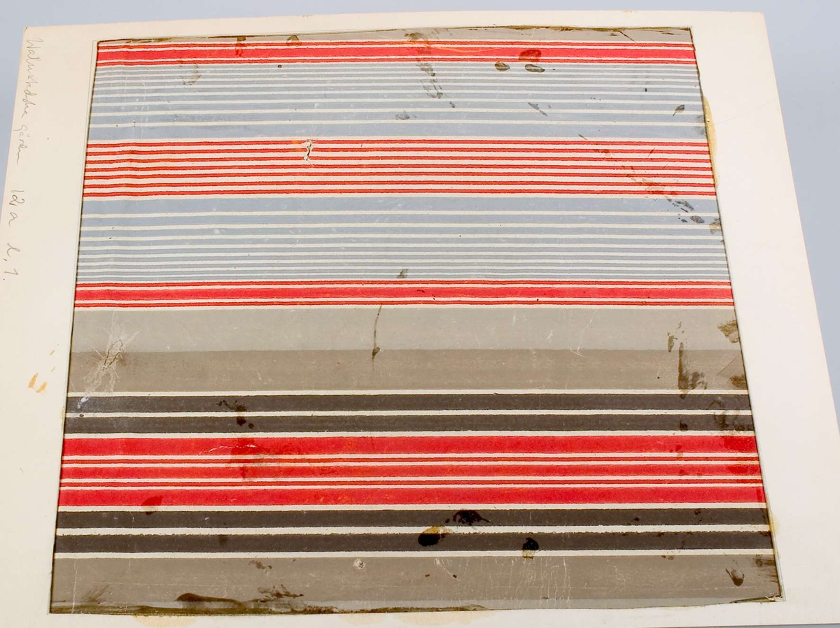 Ett tapetfragment av papper, tryckt mönster, ränder i färgerna grunt, rött, blått, och svart på vit botten. Uppklistrat på papp. Blyertstext:  "Walmstedtska gården, 12a l. 1".
