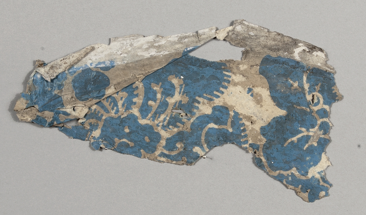 Tapetprovet består av 15 st. fragment. Grundfärgen är beige, med schablonmönster i blått. 