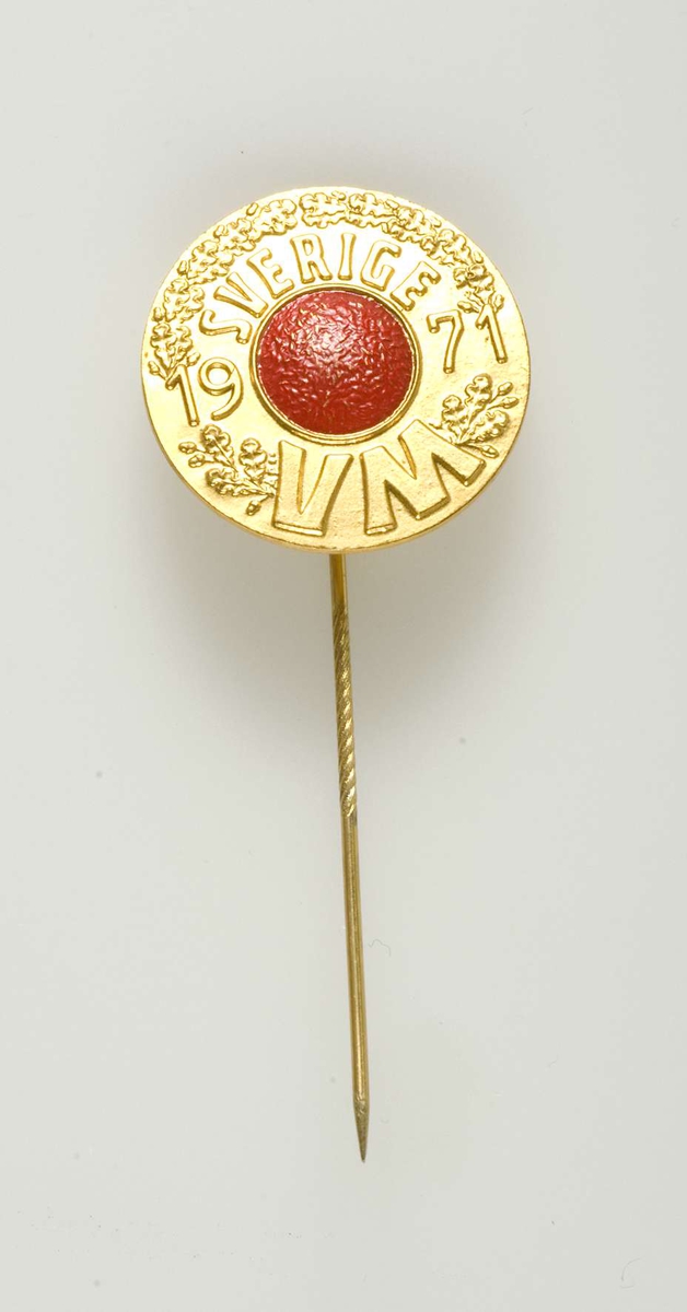 Runt märke som fästes med nål av gul metall. Text: Sverige 1971 VM. En röd bandyboll inom växtornamentik.