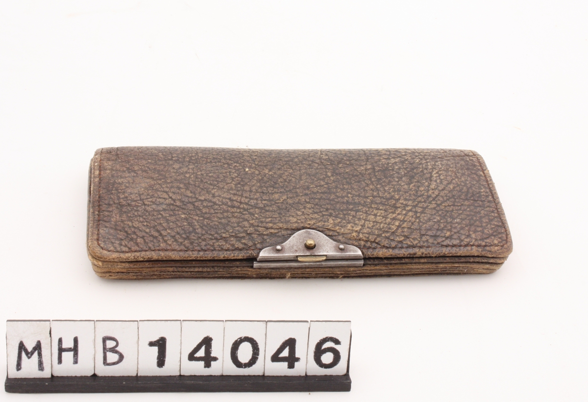 Rektangulær lommebok i lær med metallås midt på den ene langsiden. Lommeboken kan åpnes, og der er 2 rom til sedler, 1 rom til mynter med metallbeslag og påfølgende 2 rom til sedler. En nøkkel ligger i rommet til mynter.