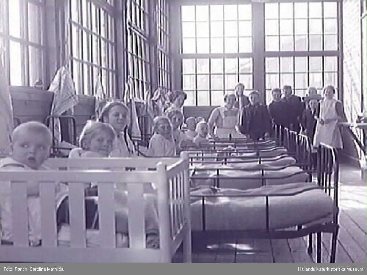 Solveranda på Apelvikens sanatorium med personal och patienter år 1915. I sängarna sitter barnpatienterna och ler mot fotografen. Närmast kameran sitter ett litet barn i spjälsäng och baktill står en grupp ungdomar, bl a en flicka klädd i kappa och vid hatt.