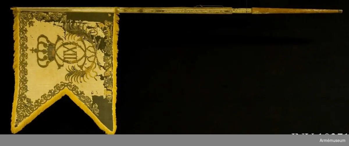 Grupp B I.
Duk av vit damast, varå målat i guld omvänt lika på båda sidor Karl XII:s namnchiffer, dubbelt C under sluten rödfodrad kunglig krona, infattat av två nedtill korsade och med rosett hopknutna, lyrformigt böjda palmkvistar. Längs kanten bård av palmkvistar och ornament liknande dubbla C samt i hörnen små öppna kronor. Längs kanten en 6 cm bred frans av gult silke. Duken fäst vid stången med två vita sidenband och förgyllda mässingsspikar. Bredd 119 och 76 cm. Stång av furu, ovan greppet med sex refflor och tre förstärkande järnskenor, målad vit, men färgen mestadels  avnött, försedd med löpande bärring och klack av järn.

Längd totalt 320 cm, till greppet 89 cm, greppet 19 cm och till duken  116 cm. Diameter upptill 3,5 cm, nedom duken 5,5 cm, ovan  greppet 6,4 cm och i greppet 5 cm. Klacken 4 cm hög och holken  6 cm. Spets av förgylld mässing.