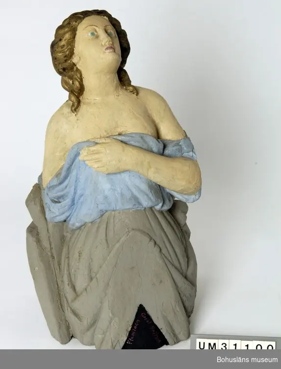 Föremålet visas i basutställningen Kustland,  Bohusläns museum, Uddevalla.

Galjonsfigur från mitten av 1800-talet föreställande kvinnofigur iförd en löst draperad klädnad som bildar böljande veck.  Kvinnan blickar mot skyn, har en uppsatt frisyr med stor knut i nacken och nedfallande lockar vid öronen, nakna axlar och hon håller klänningens överdel med vänster hand över bröstet. Baksidan är  delvis obearbetad.
Figuren är skuren ur ett stycke trä, krederad och bemålad. 
Nuvarande bemålning är med största sannolikhet utförd av C G Bernhardson med håret i guld, klänningens överdel ljusblått, nederdelen grå, baksidan och en del av framsidans nederdel svart.
Nertill på galjonsbildens framsida har Bernhardson skrivit med röda bokstäver: 
"Flamingo Great Britain", troligen namnet på ett fartyg, kanske det som galjonsbilden en gång hört till. Uppgifter saknas om fyndomständigheter.
Upphängningsögla på baksidan med nylonsnöre.
Torrsprickor, färgbortfall som målats över.