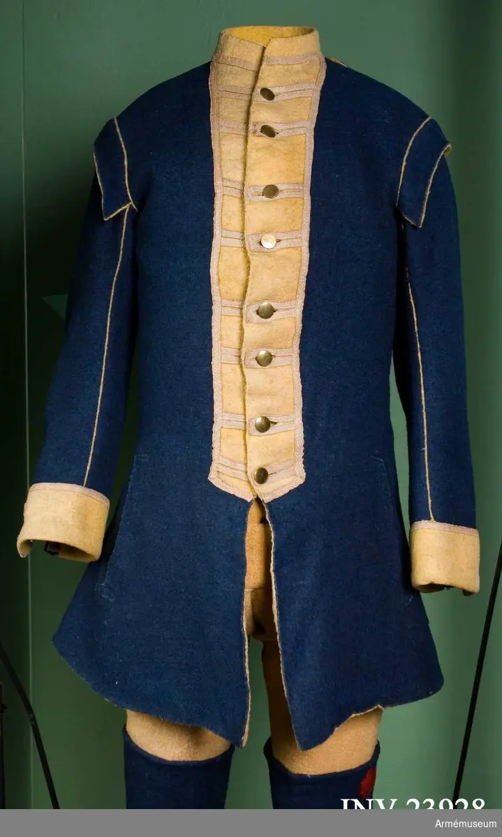 Jacka av blått kläde med gula bröstrevärer, krage och ärmuppslag, fodrad med gul boj samt linne i ärmarna. Försedd med mässingsknappar och vävda band som dekoration.
