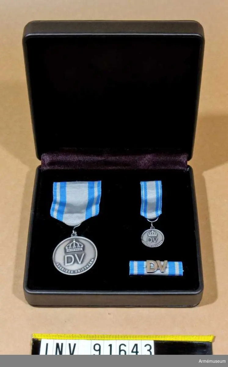 Etui innehållande medalj i silver, minimedalj samt släpspänne. 
På medaljernas framsida finns text samt en kunglig krona. På baksidan endast text.
Band i blått med brett grått streck i mitten och ett grått streck på vardera sidan.