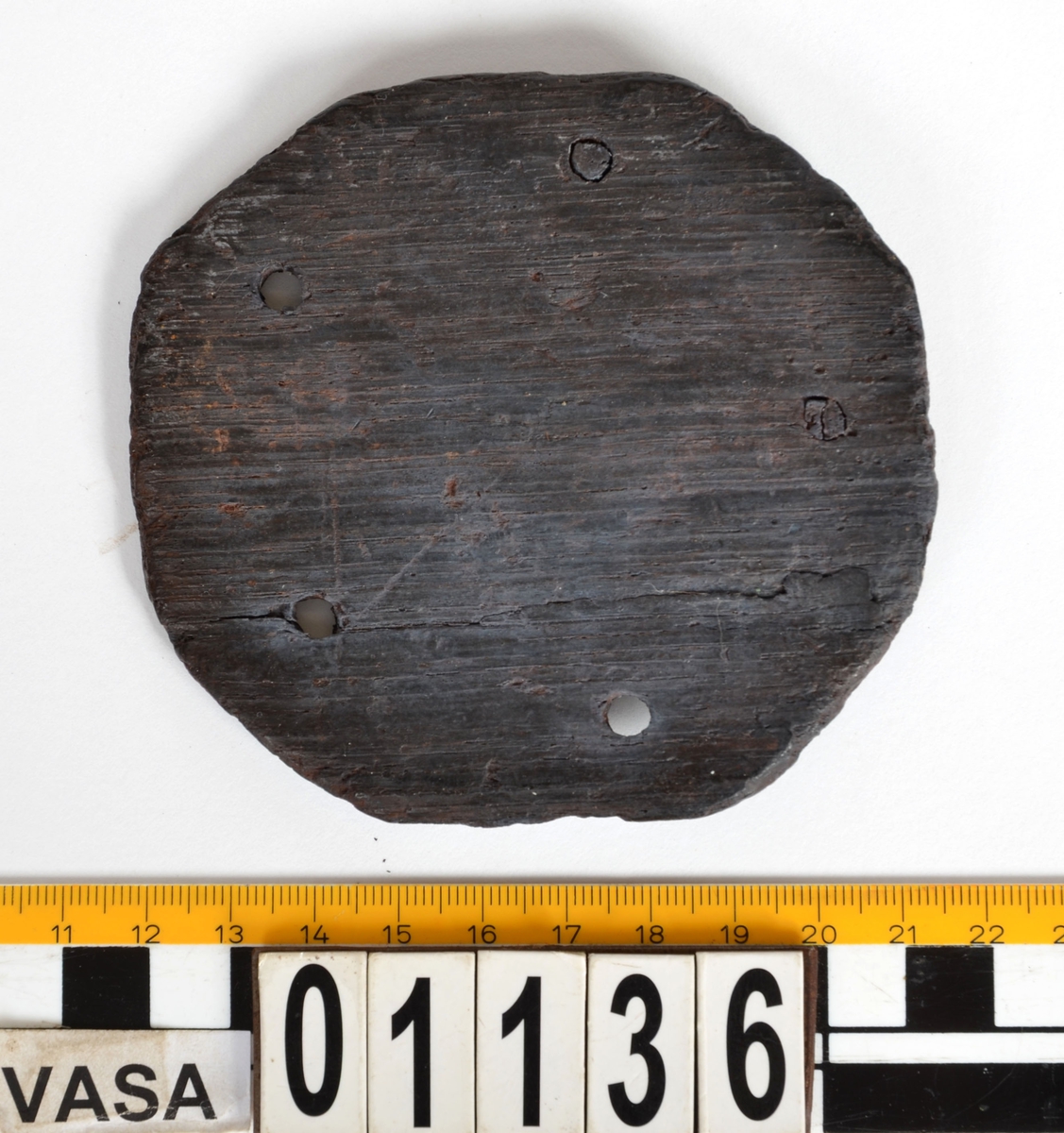 Platta till ett timglas. Åttasidig träplatta med fem genomgående hål runtom. Rest av pinne i ett hål.

Text in English: Eight-sided wooden plate identified  as the end cap for a sand glass (hourglass), the most common type of time-keeping device on board ships until the 19th century. The size is correct for a half-hour glass, which was used to time the watches and the length of a man?s ?trick? as steersman before he was relieved. Vasa carried at least two sandglasses, but probably many more, since they were easily broken.