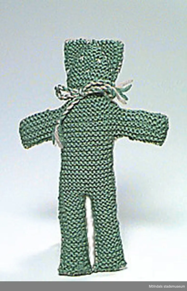 En docka i form av en nalle i grönt och grått. Ull eller konstfibrer.Undervisningsmaterial för åk 4 och 5 på Kvarnbyskolan. Se även 02645-02652. Övriga uppgifter se 02643:1.
