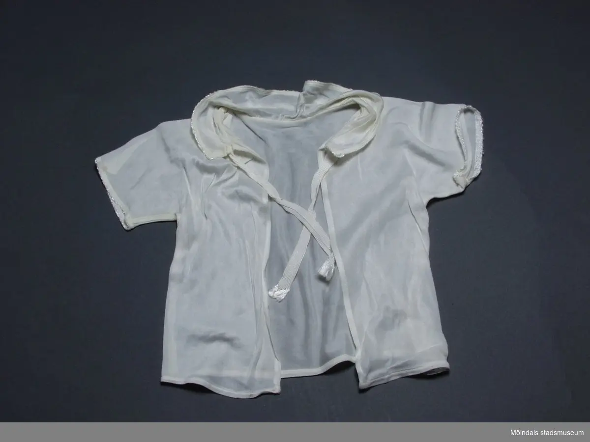 Spädbarnsskjorta i vit charmeuse. Halsrysch och ärmar är kantade med en liten langett.
