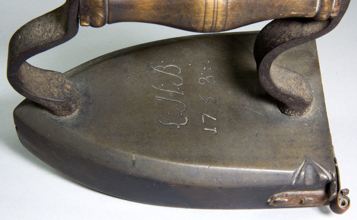 Lodstrykjärn av mässing. Handtag svarvat av trä, på två svängda ståndare av gjutjärn. Ovansidan av järnet en ristad signatur: "L.H.B 1758". Baktill på järnet en öppningsbar lodlucka, som låses med fällbar hasp.