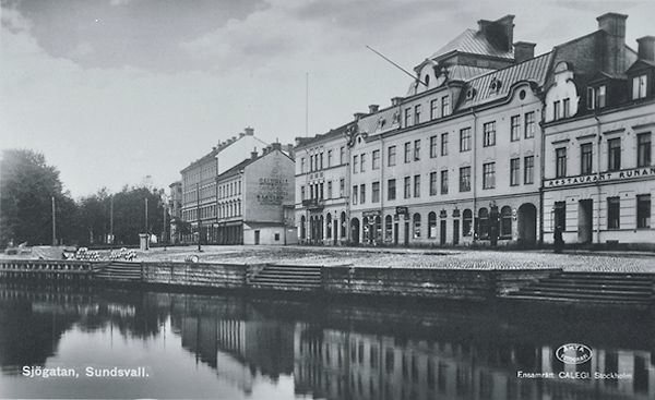 Till vänster Selångersån till höger Sjögatan med bebyggelse. På fastigheten längst till höger syns texten "Restaurant Runan". Bildtext på vykort "Sjögatan, Sundsvall".