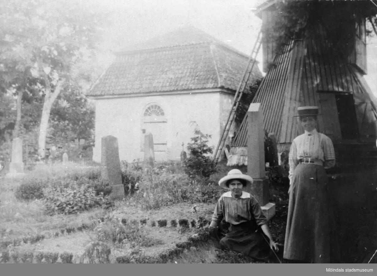Valdeborg Johansson (sittandes) och mostern Josefina Eriksson framför klockstapeln på Kållereds kyrkogård cirka 1910. De är vid gårdsgraven till Vommedal Östergård, "Olas".