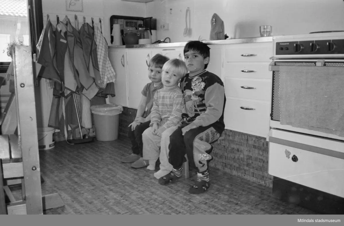 Tre barn som sitter på en liten pall framför en diskbänk. De tittar mot fotografen. Katrinebergs daghem, 1992-93.