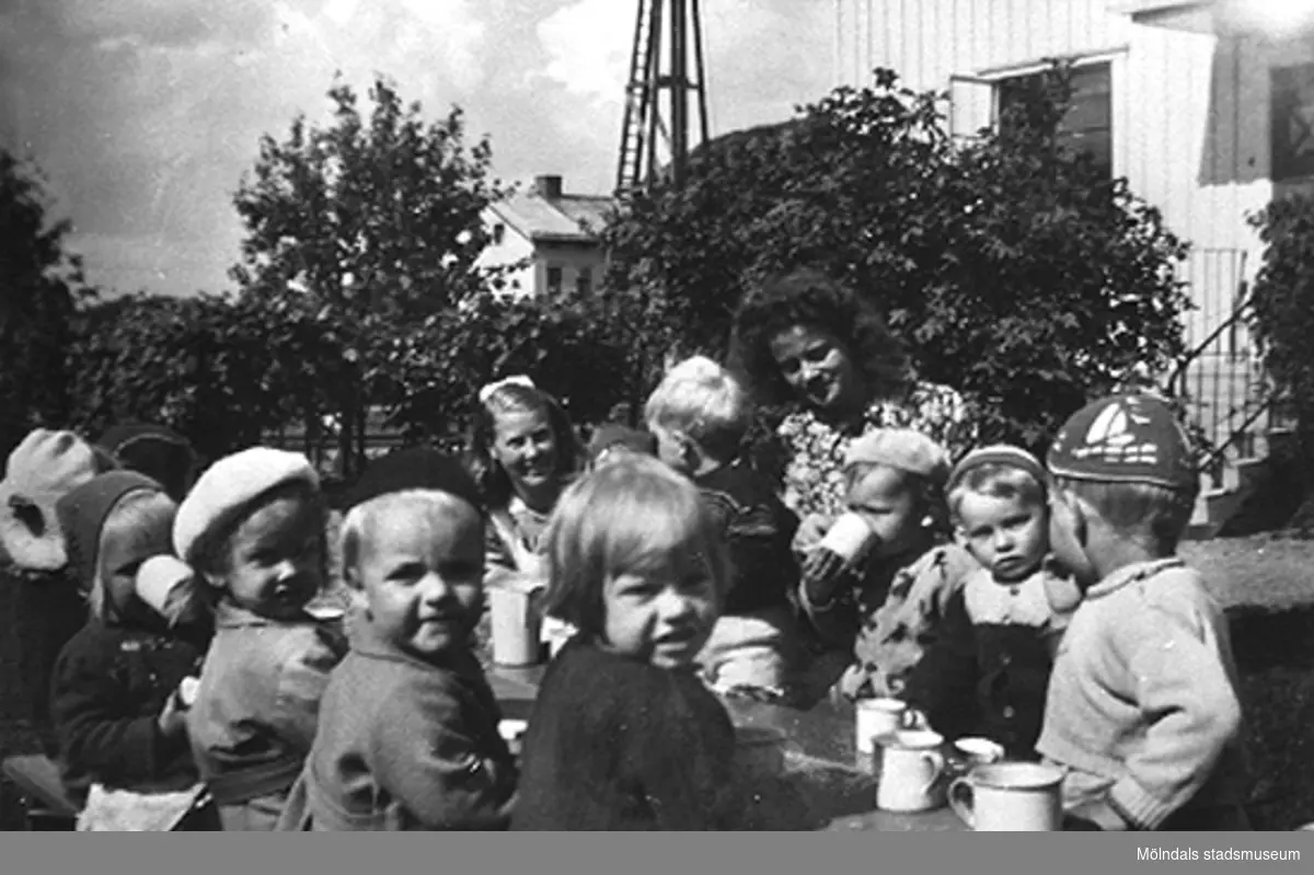 Lärare och barn fikar i trädgården vid 
Krokslätts daghem, Dalhemsgatan 7 i Krokslätt. Praktikant Margit Emilsson (gift Wannerberg -52) står vid barnen. 
Under Margits praktik som biträde 1945-10-01 - 1946-08-01.