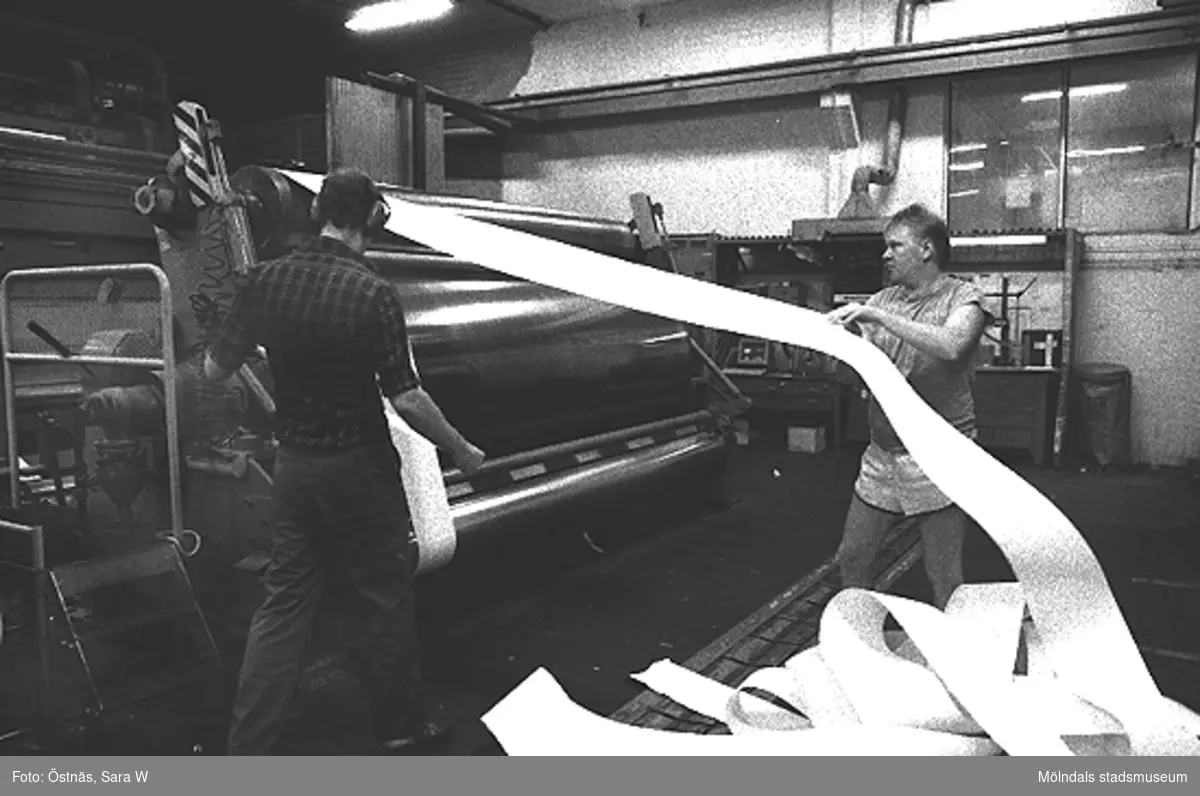 Två män vid en maskin för papperstillverkning, 1980-tal. Mannen som drar i papperet är Kent-Erik Pettersson.
Bilden ingår i serie från produktion och interiör på pappersindustrin Papyrus.
