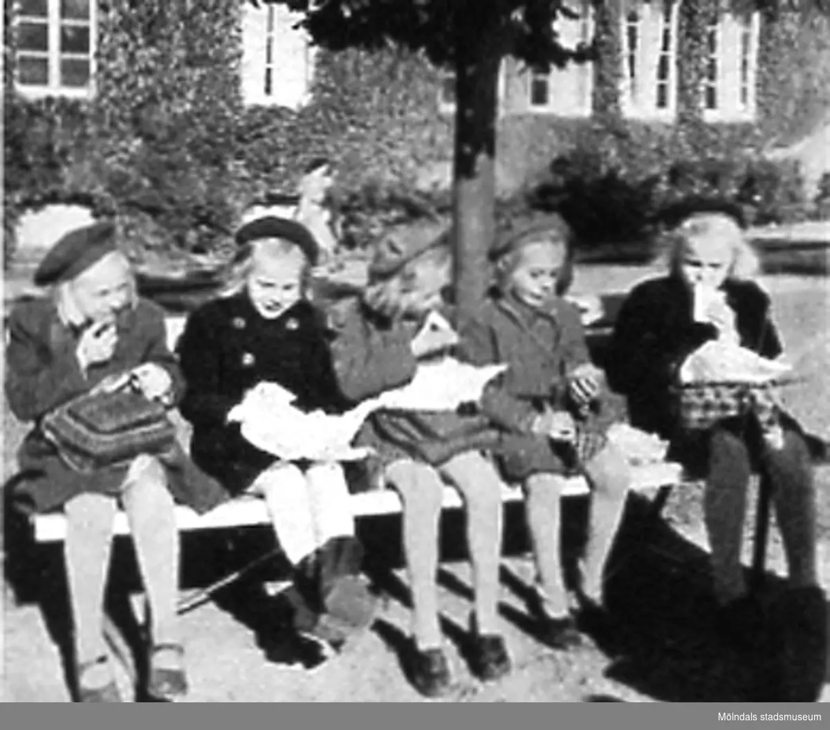 Fem flickor sitter på en bänk och har matrast utanför Kvarnbyskolan (Centralskolan), okänt årtal. Avfotograferad ur "Mölndal 1922 - 1947".