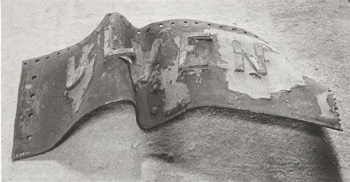 Fartygsplåt med ubåten ULVENS namn i metallbokståver. Plåten tillbucklad i samband med minexplosionen.