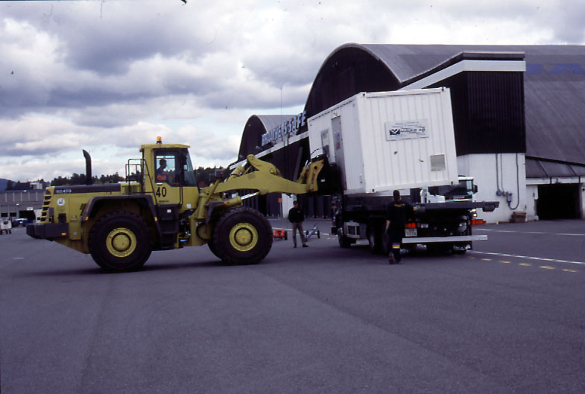 Lufthavn, 1 person i 1 kjøretøy løfter 1 container opp på lasteplanet til 1 lastebil.  1 person og 1 hangarbygning til Braathens SAFE i bakgrunnen.