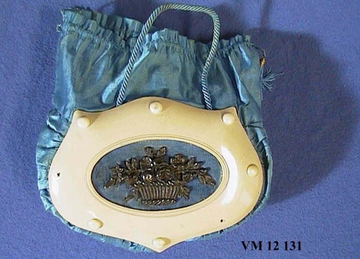 Väska, blå satin i påsform. På nedre delen påsatt en vit celluloidram i vars mitt en metallblomkorg är fästad på blå sammet.