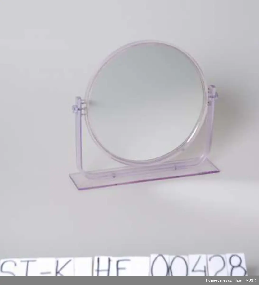 Rundt speil med ramme og stativ av transparent, lys lilla plast. Speilet kan snurres rundt i rammen, og det er speil på begge sidene.