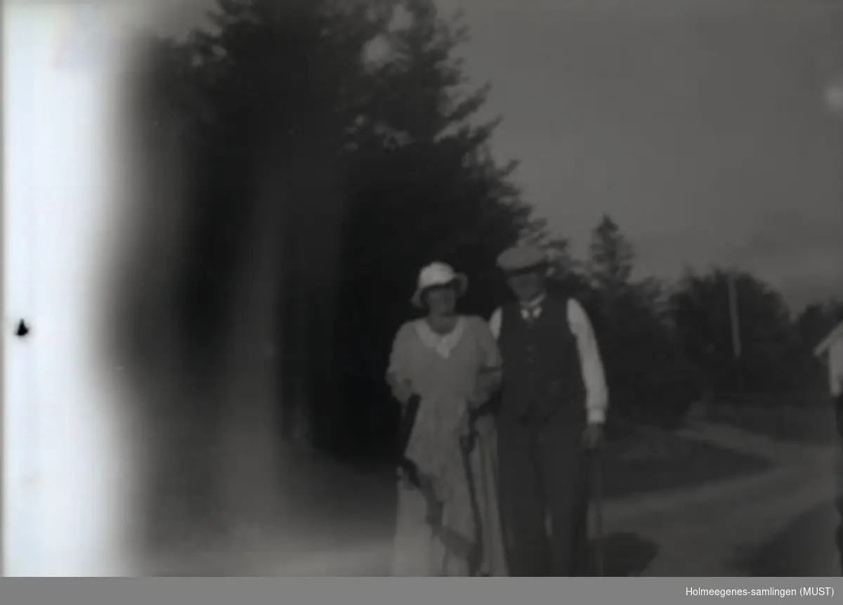 En kvinne i kjole og en mann i skjorte og vest, begge har hatt på. Utendørs i fint vær. Noe skog i bakgrunnen.