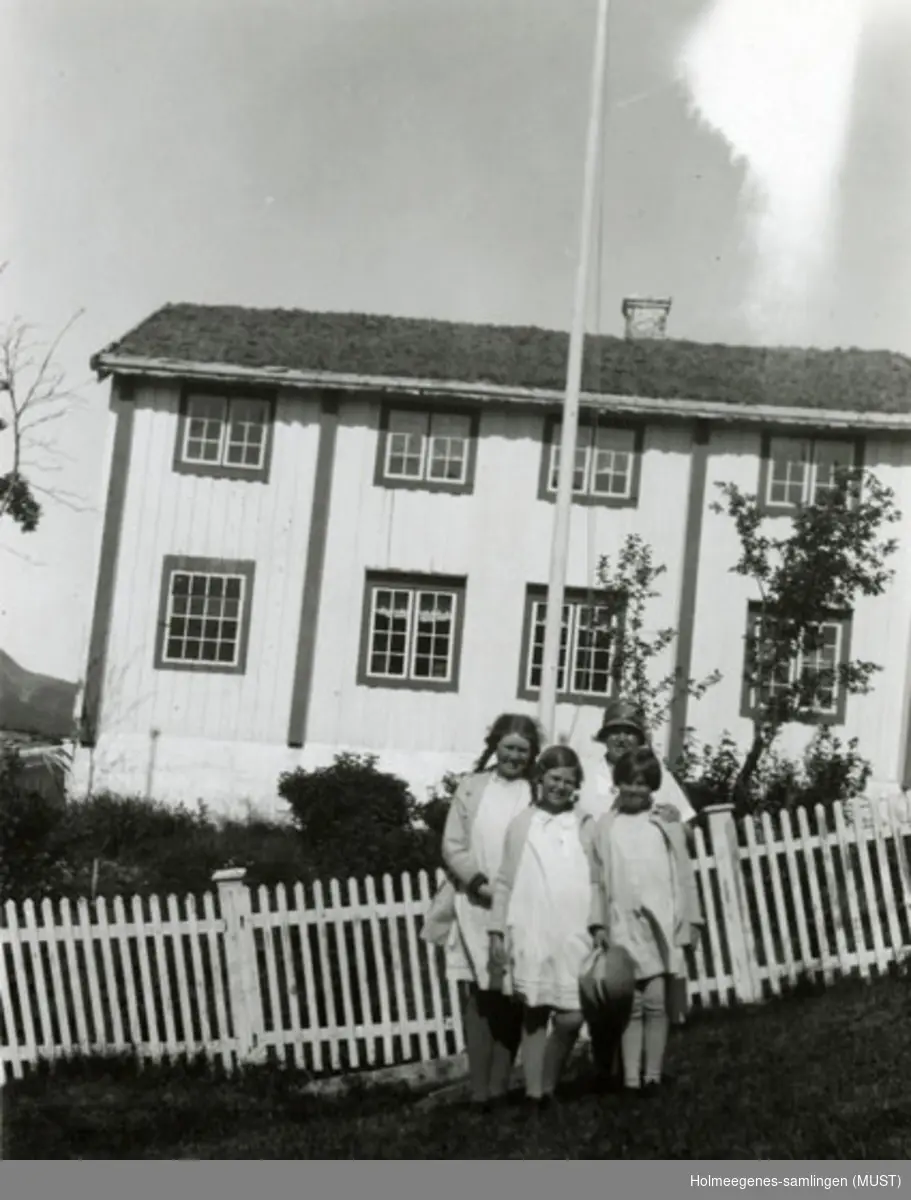 En kvinne og tre jenter i sommerkjoler står foran et hvitmalt tregjerde, i bakgrunnen et bolighus med to etasjer. Se også ST.K.HE 2007-011-0111 til -0126.
