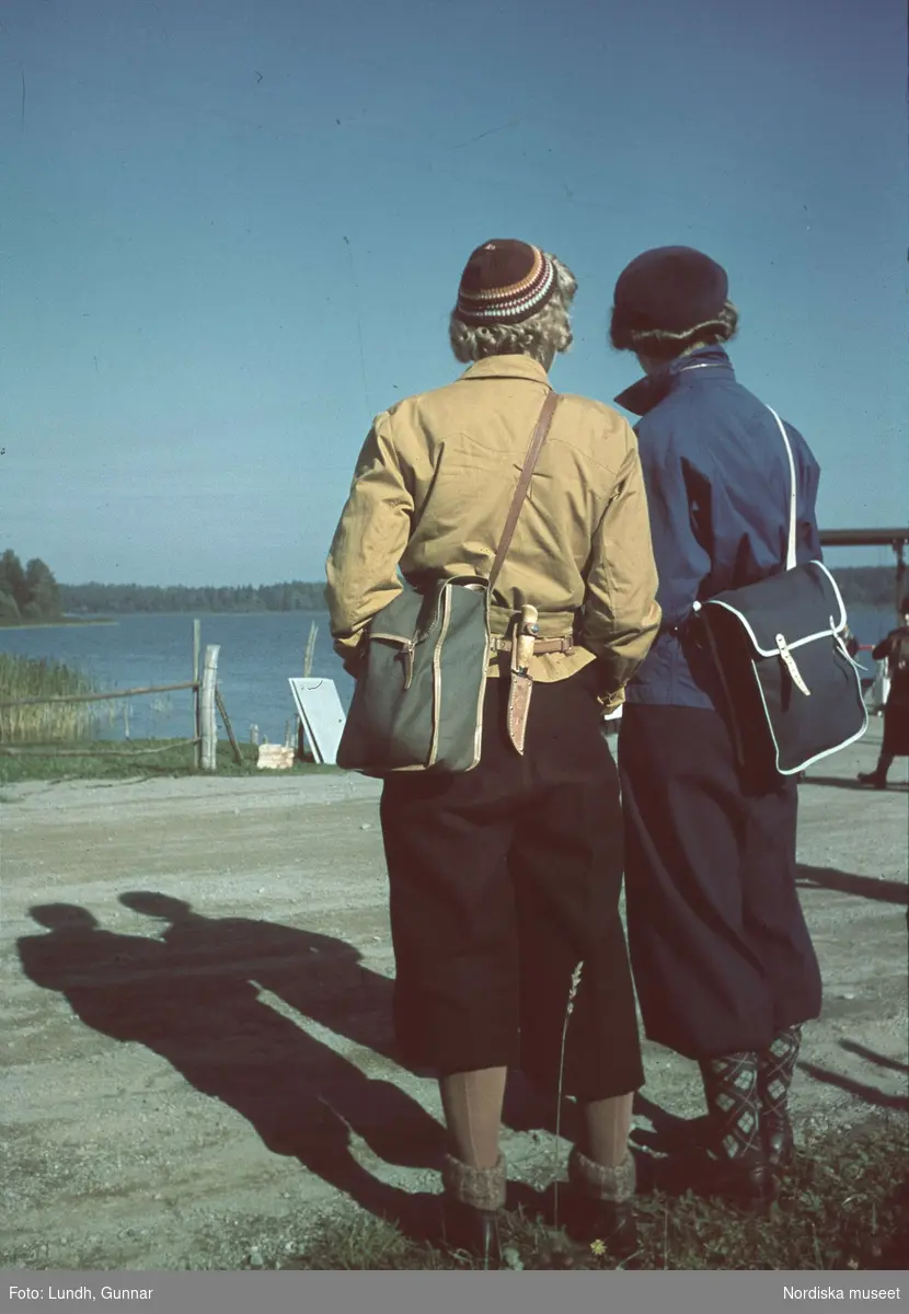 Två kvinnor i fritidskläder och med axelremsväskor står med ryggarna vända mot kameran och tittar ut över vatten.